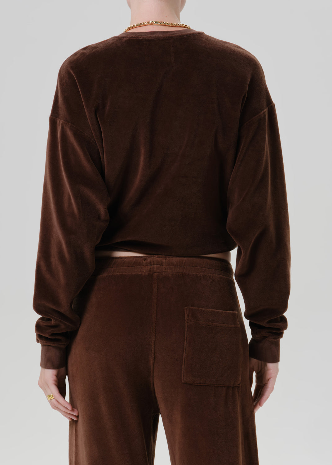 Woman wearing a dark brown crewneck sweatshirt with dark brown wide leg pants viewed from the back on a grey background.