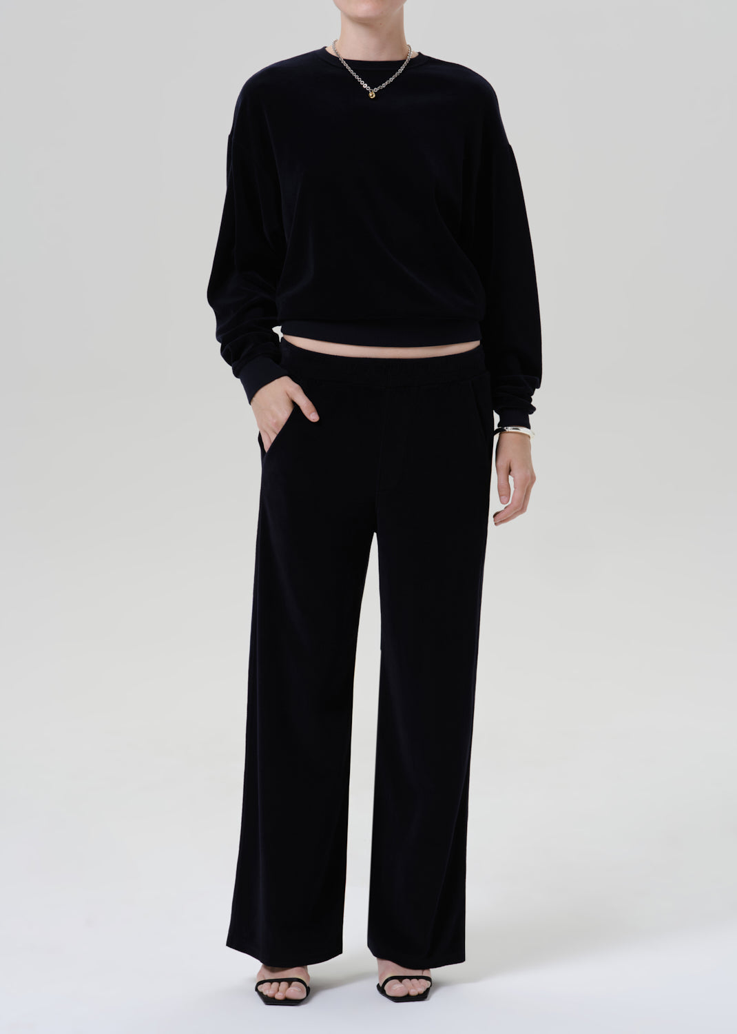 A model wearing a black long-sleeve cropped sweatshirt and matching black wide-leg trousers, styled with a delicate gold chain necklace and black flat sandals against a grey background.