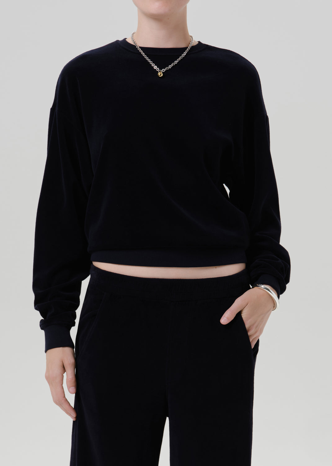 A model wearing a black long-sleeve cropped sweatshirt and matching black pants, accessorized with a gold chain necklace and bracelet, against a grey background.