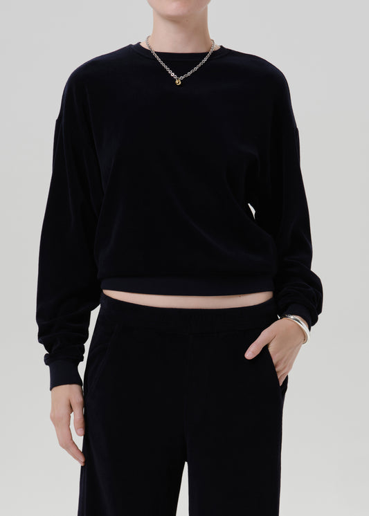 A model wearing a black long-sleeve cropped sweatshirt and matching black pants, accessorized with a gold chain necklace and bracelet, against a grey background.