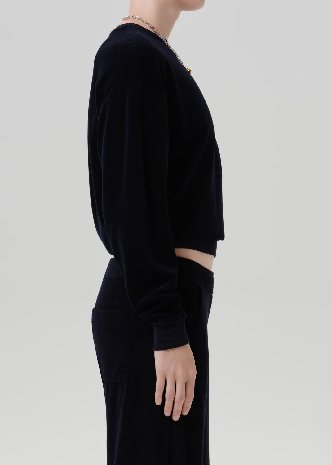 A side-profile view of a model wearing a black velvet long-sleeve crop top and matching black velvet trousers against a grey background.