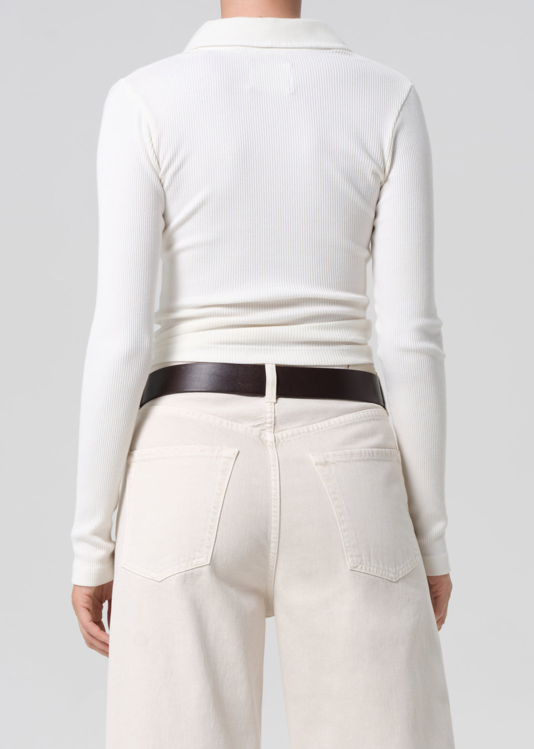 Person wearing a white long-sleeve top and cream pants with a brown belt on a light gray background