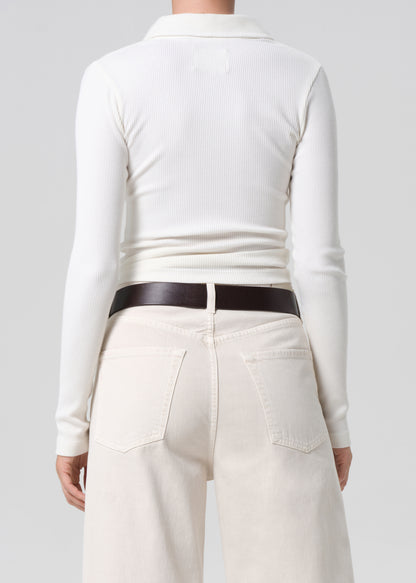 Person wearing a white long-sleeve top and cream pants with a brown belt on a light gray background