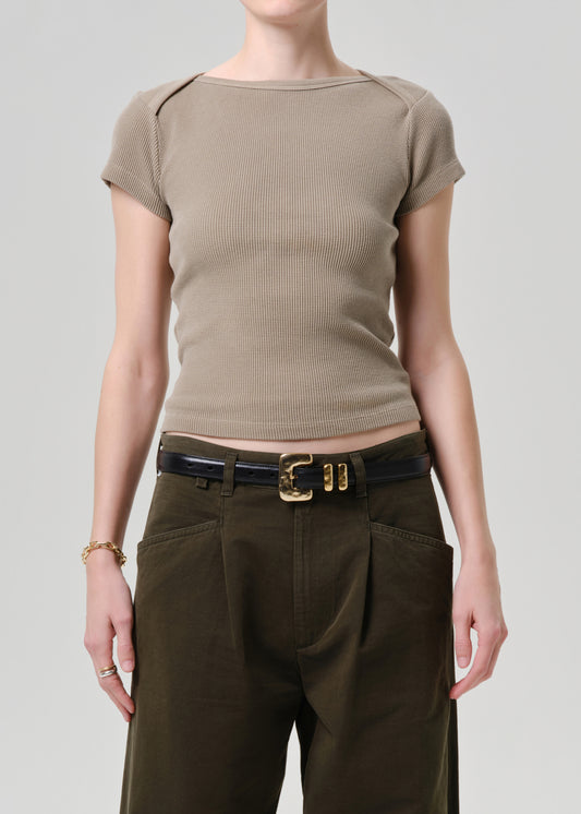A model wearing a beige short-sleeve ribbed knit top with shoulder detailing, tucked into dark olive green pleated trousers with a black belt featuring a gold buckle.