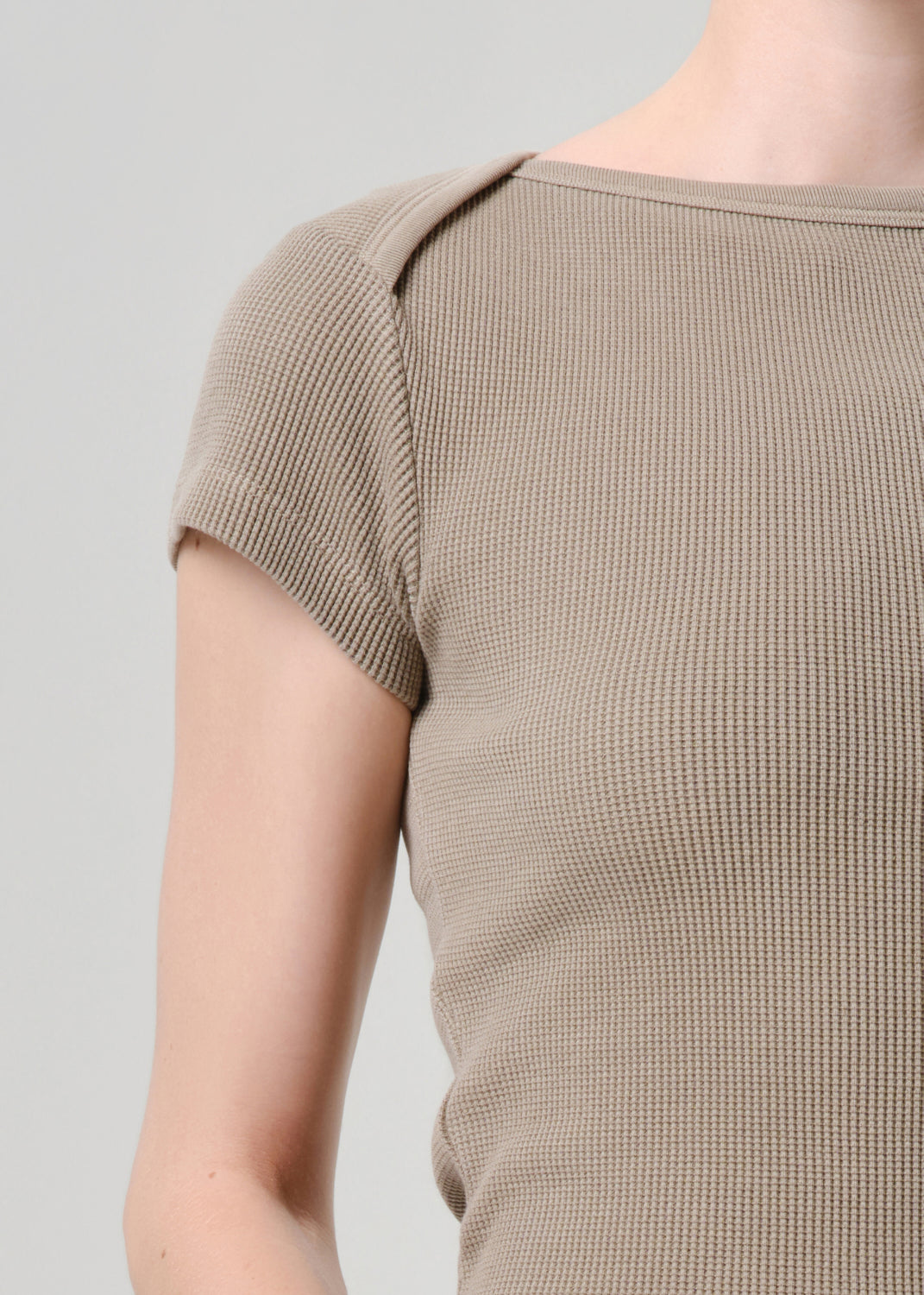 Close-up of a person wearing a short-sleeve ribbed knit top in a warm taupe-beige color against a grey background.