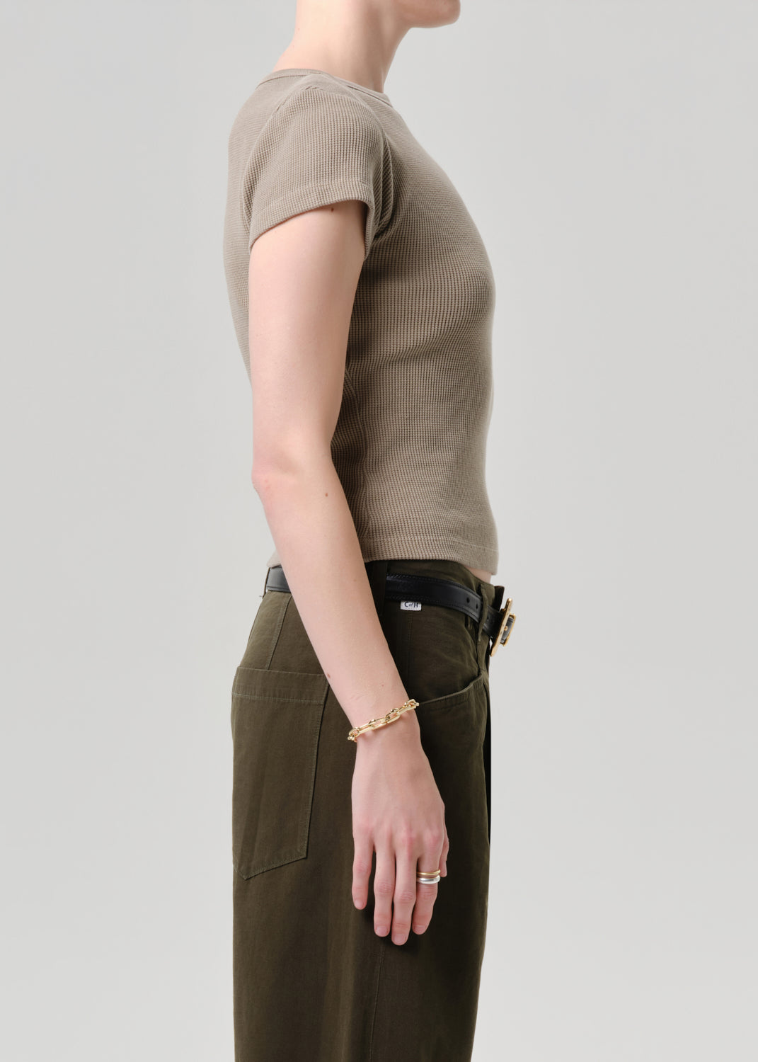 Side profile of a model wearing a short-sleeve ribbed knit top in taupe and olive green trousers, accessorized with a gold chain bracelet and ring against a grey background.