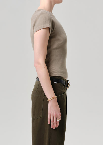 Side profile of a model wearing a short-sleeve ribbed knit top in taupe and olive green trousers, accessorized with a gold chain bracelet and ring against a grey background.