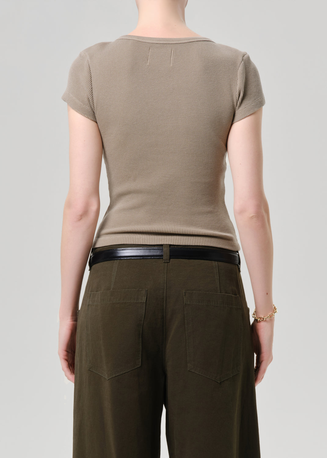 Rear view of a model wearing a taupe short-sleeve ribbed knit top and dark olive green wide-leg trousers with a black belt, against a grey background.