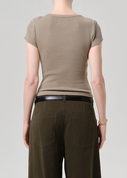 Rear view of a model wearing a taupe short-sleeve ribbed knit top and dark olive green wide-leg trousers with a black belt, against a grey background.