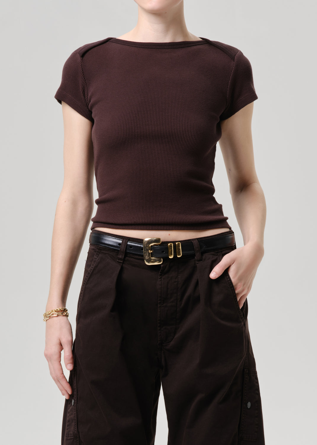 A model wearing a dark brown ribbed short-sleeve boat-neck crop top and dark brown cargo pants with a black belt, photographed against a grey background.