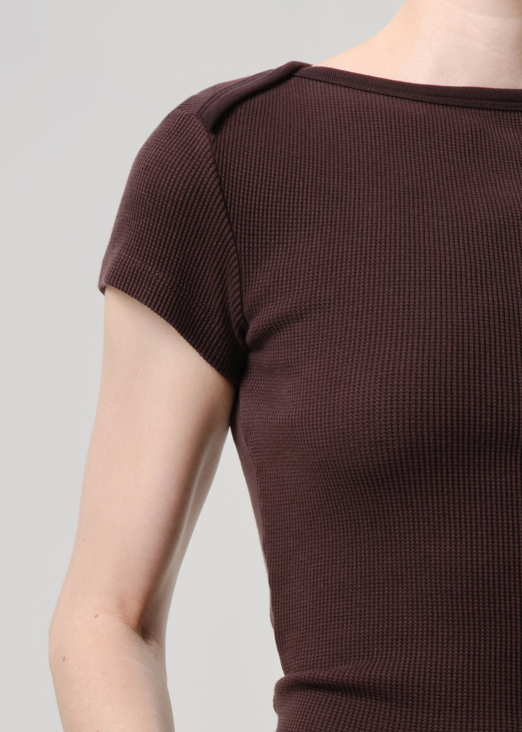 Close-up of a person wearing a short-sleeve ribbed knit top in a deep plum-brown color against a grey background.