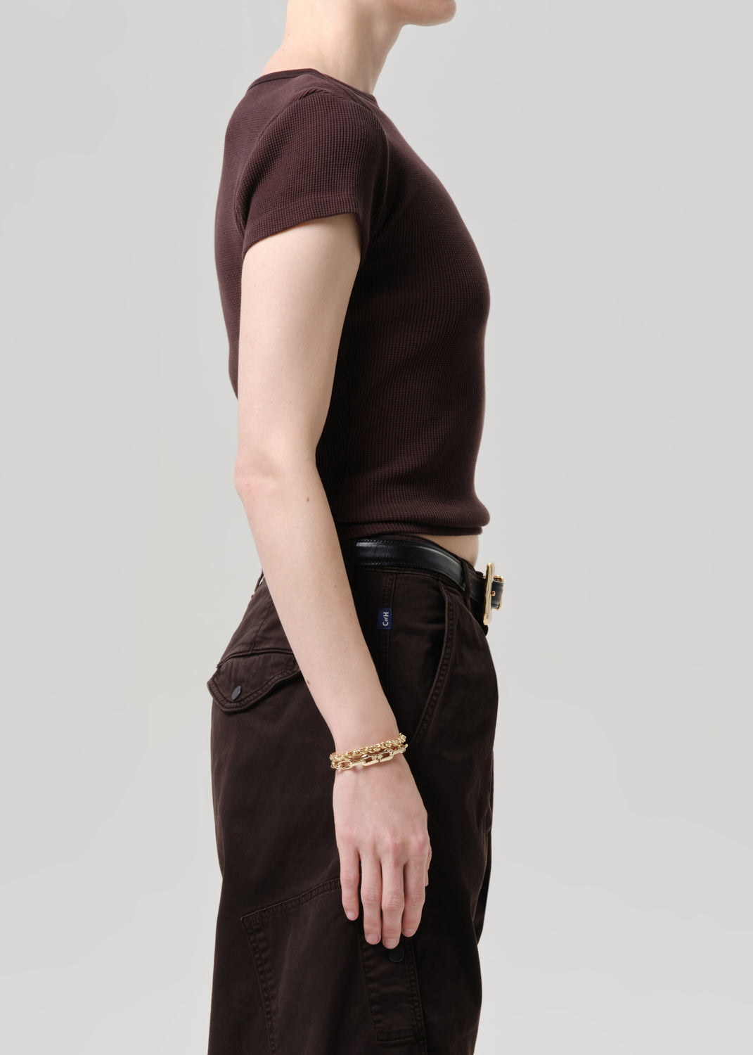Side profile of a model wearing a dark brown short-sleeve crewneck t-shirt tucked into dark brown cargo pants with a black belt, against a grey background.