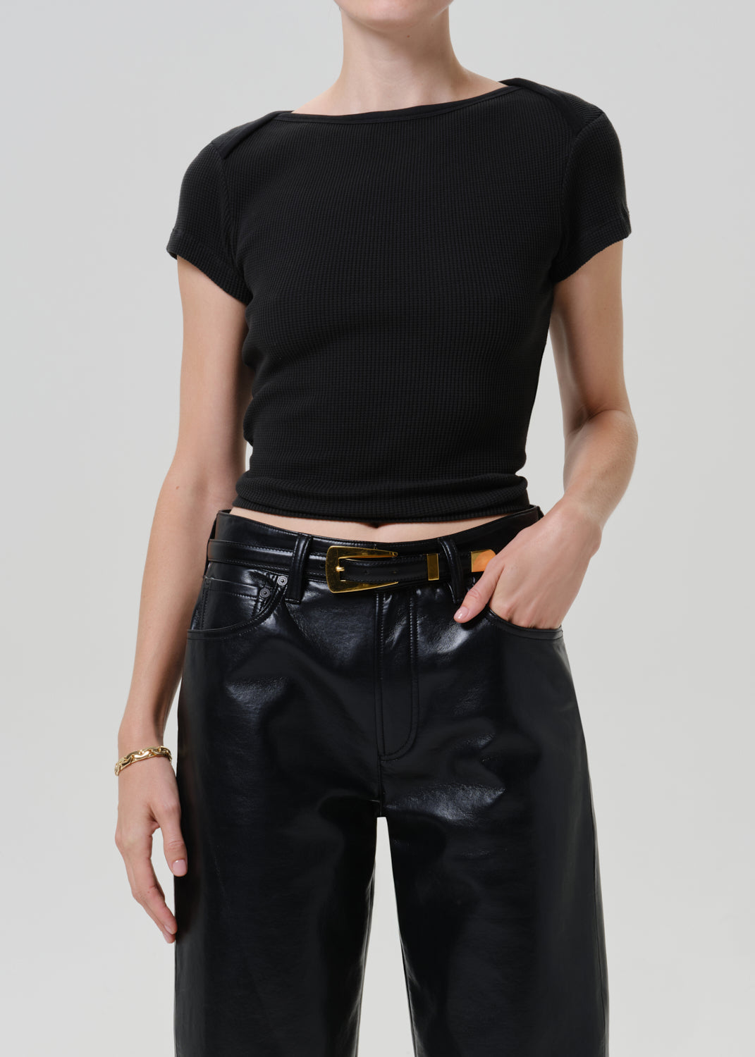 A model wearing a black short-sleeve ribbed knit top with a boat neckline, paired with black faux-leather pants and a black belt with a gold buckle, against a light gray studio background.