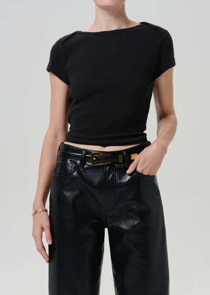 A model wearing a black short-sleeve ribbed knit top with a boat neckline, paired with black faux-leather pants and a black belt with a gold buckle, against a light gray studio background.