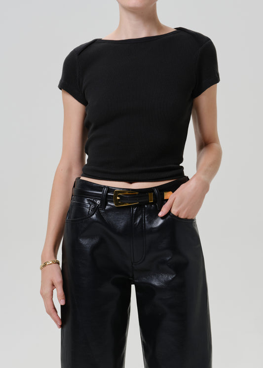 A model wearing a black short-sleeve ribbed knit top with a boat neckline, paired with black faux-leather pants and a black belt with a gold buckle, against a light gray studio background.