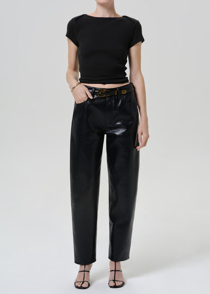 A model wearing a black short-sleeve crop top, black high-waisted faux-leather trousers with a brown belt, and black strappy heels, photographed against a light gray studio background.