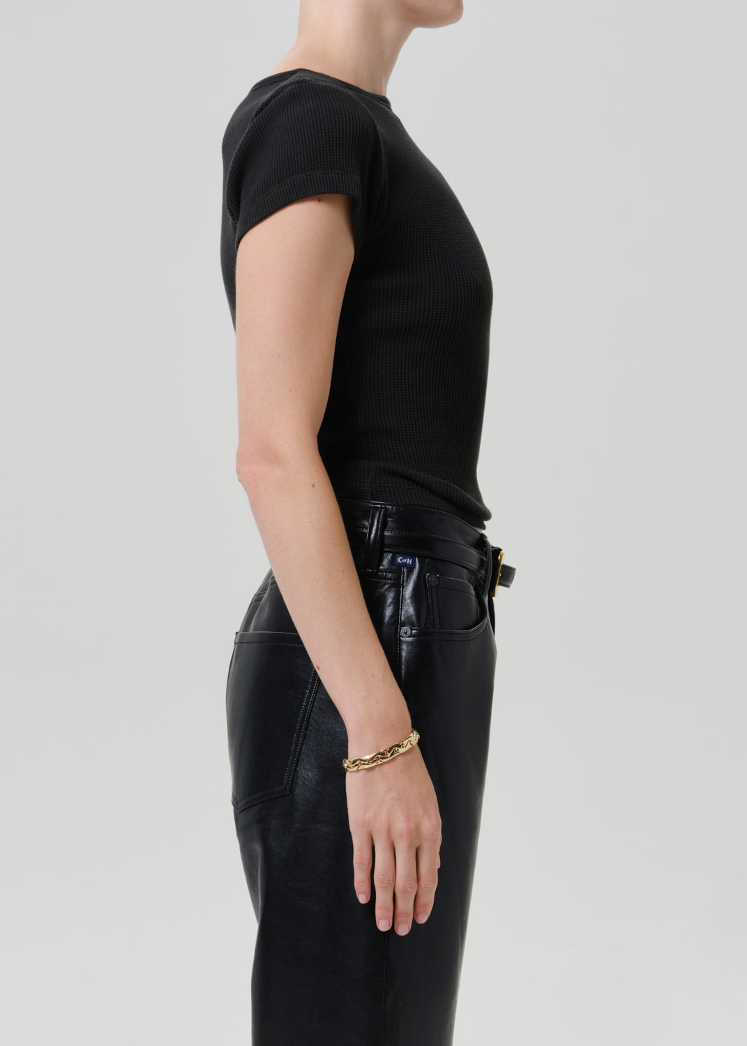 Side profile of a model wearing a black short-sleeve ribbed knit top and black leather pants against a light gray studio background.