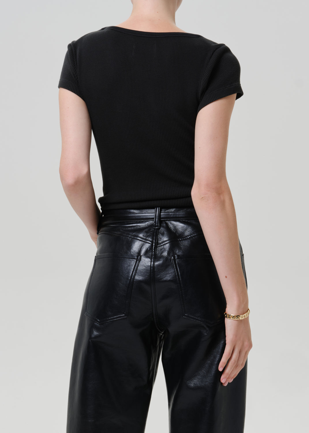 Rear view of a model wearing a black short-sleeve top and shiny black faux-leather pants against a light gray studio background.