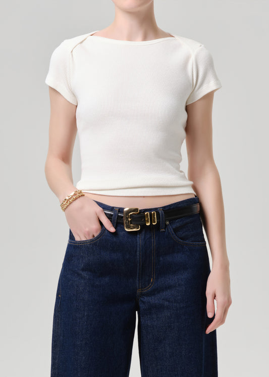 A model wearing a ribbed short-sleeve cream top and dark blue jeans with a gold belt, shown from the neck down against a grey background.