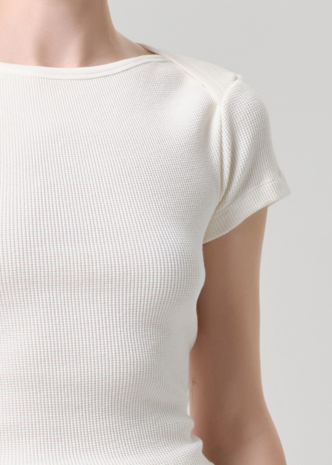 Close-up of a person wearing a short-sleeve waffle-knit top in off-white or cream, showing the textured fabric and shoulder area against a grey background.