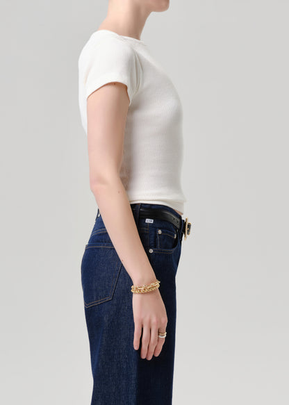 Side profile of a model wearing a white short-sleeve ribbed knit top tucked into dark blue jeans with a black belt, accessorized with a gold chain bracelet and ring, against a grey background.