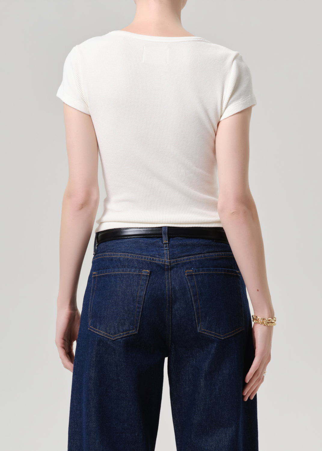 A model shown from the back wearing a fitted off-white short-sleeve t-shirt and dark blue wide-leg jeans with a black belt, against a grey background.