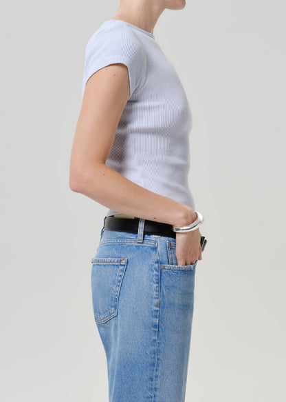 A side-profile view of a model wearing a light gray ribbed short-sleeve top, light blue jeans, and a black belt against a plain light gray studio background.