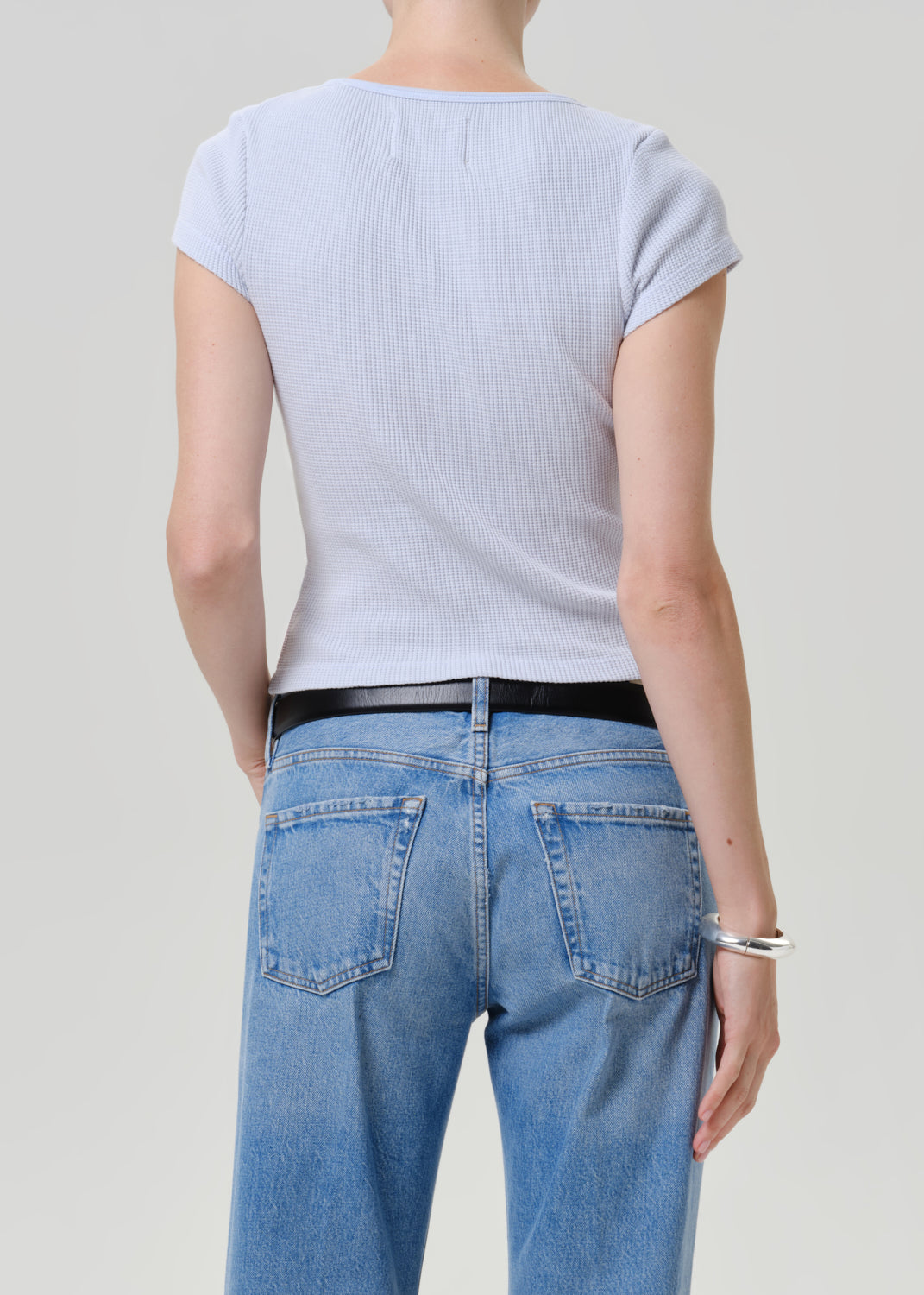 Rear view of a model wearing a light blue ribbed short-sleeve top and light blue jeans with a black belt, against a light gray studio background.