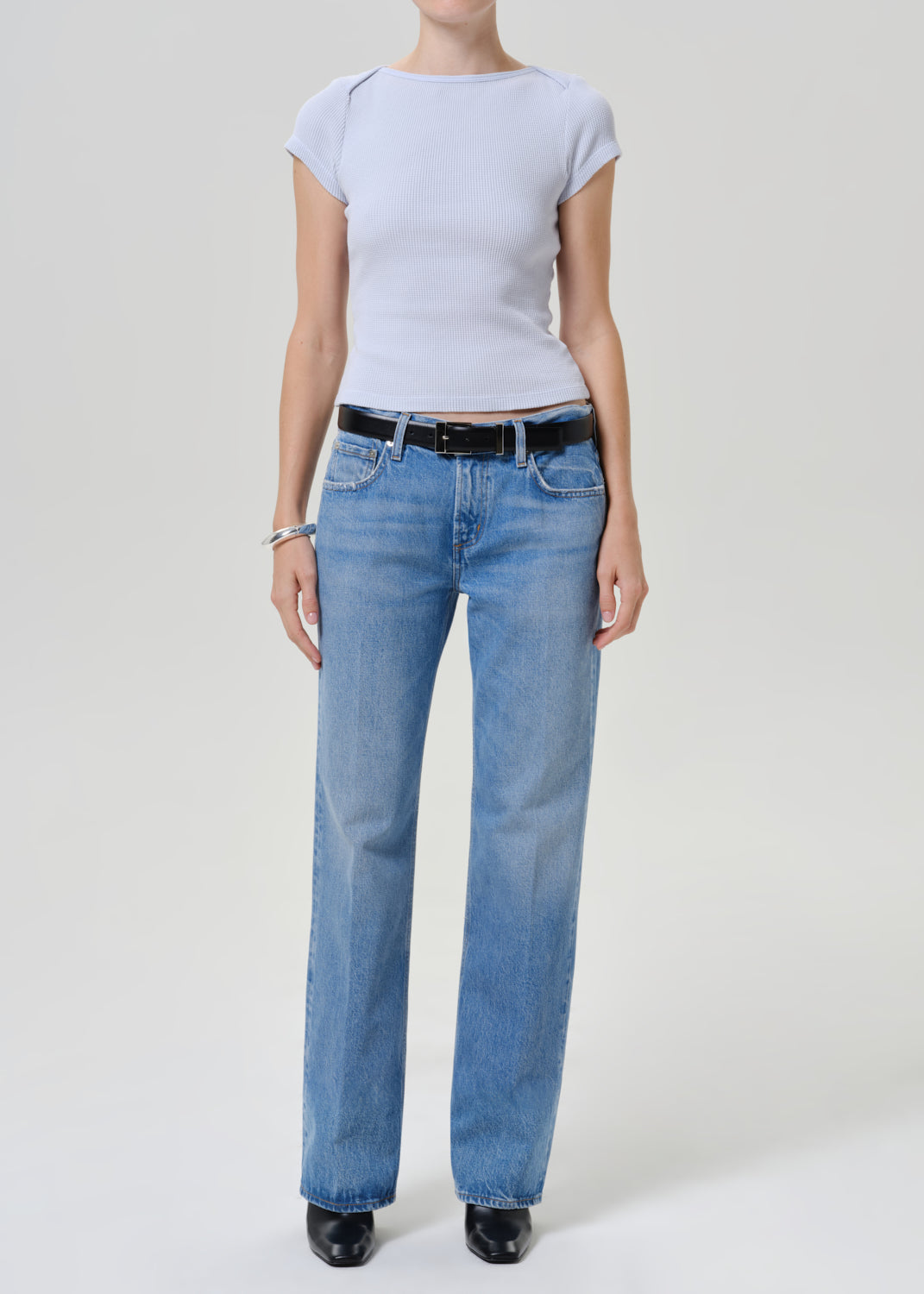 A model wearing a light blue ribbed short-sleeve top, light-wash blue straight-leg jeans with a black leather belt, and black shoes, photographed against a neutral light gray studio background.