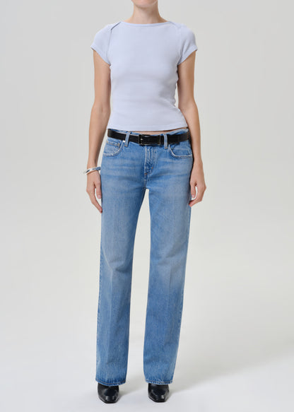 A model wearing a light blue ribbed short-sleeve top, light-wash blue straight-leg jeans with a black leather belt, and black shoes, photographed against a neutral light gray studio background.
