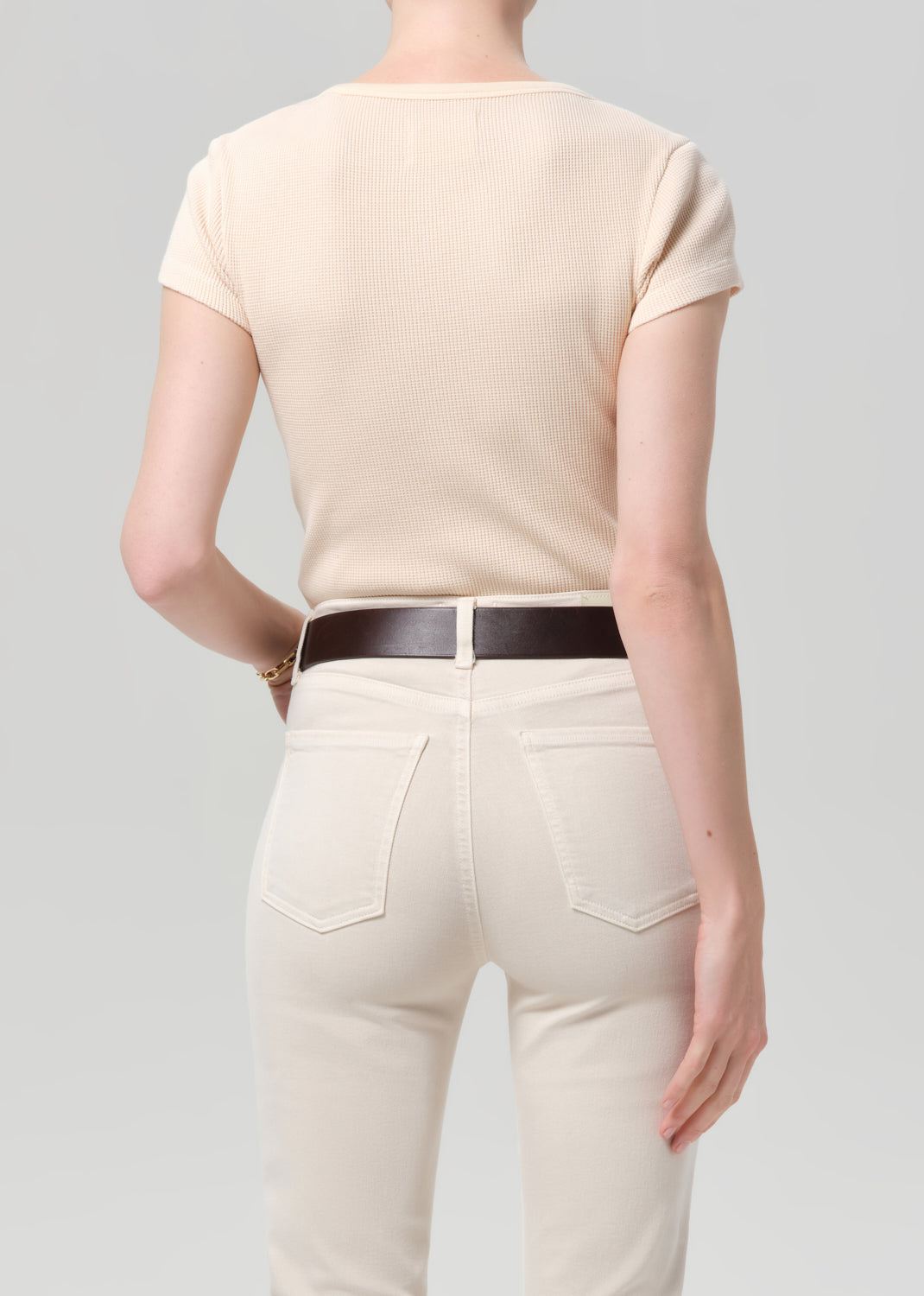 Person wearing a light peach top and cream capri pants with a brown belt on a gray background