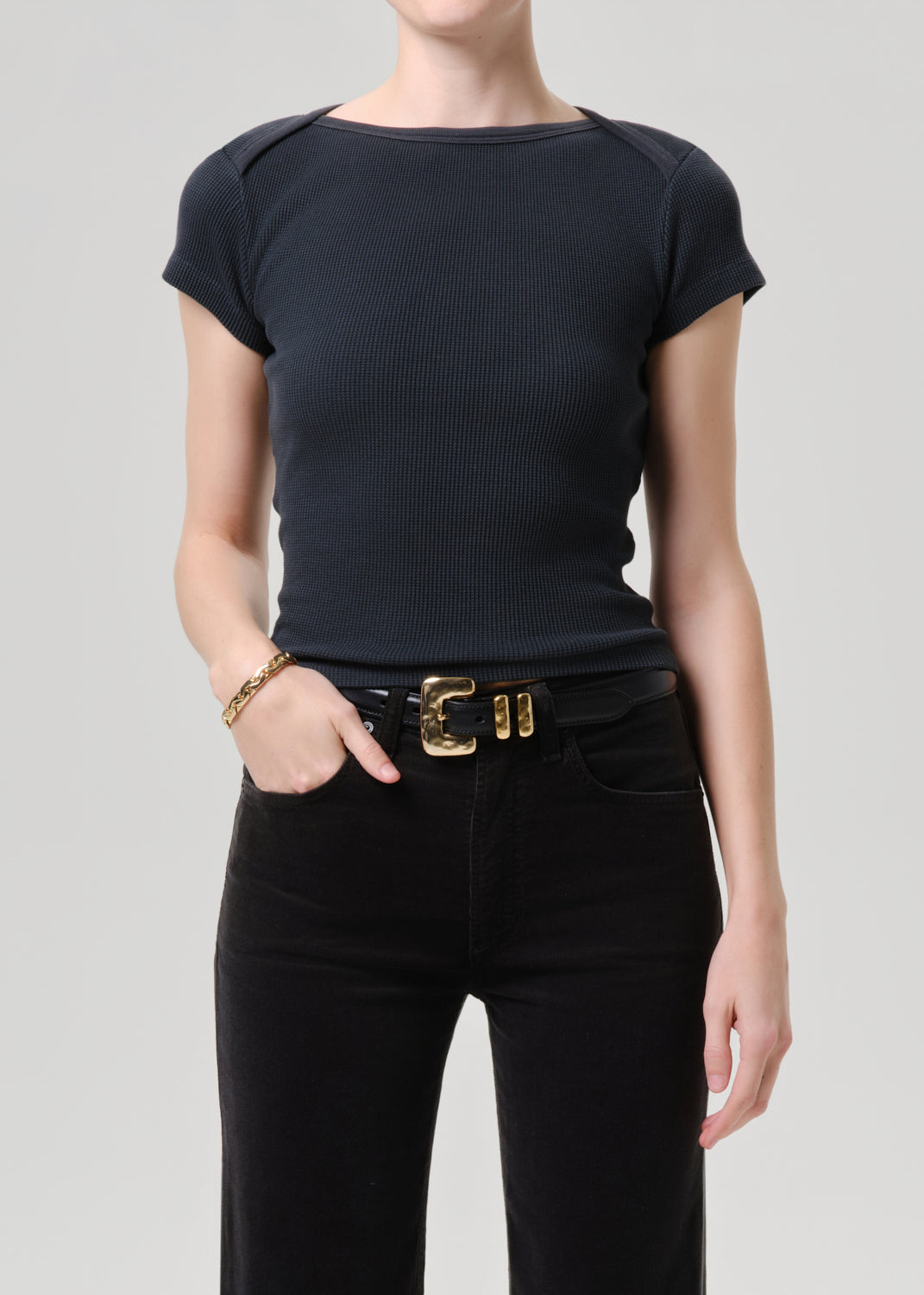 A model wearing a dark navy ribbed short-sleeve boat-neck top tucked into black jeans with a gold buckle belt, shot against a grey background.