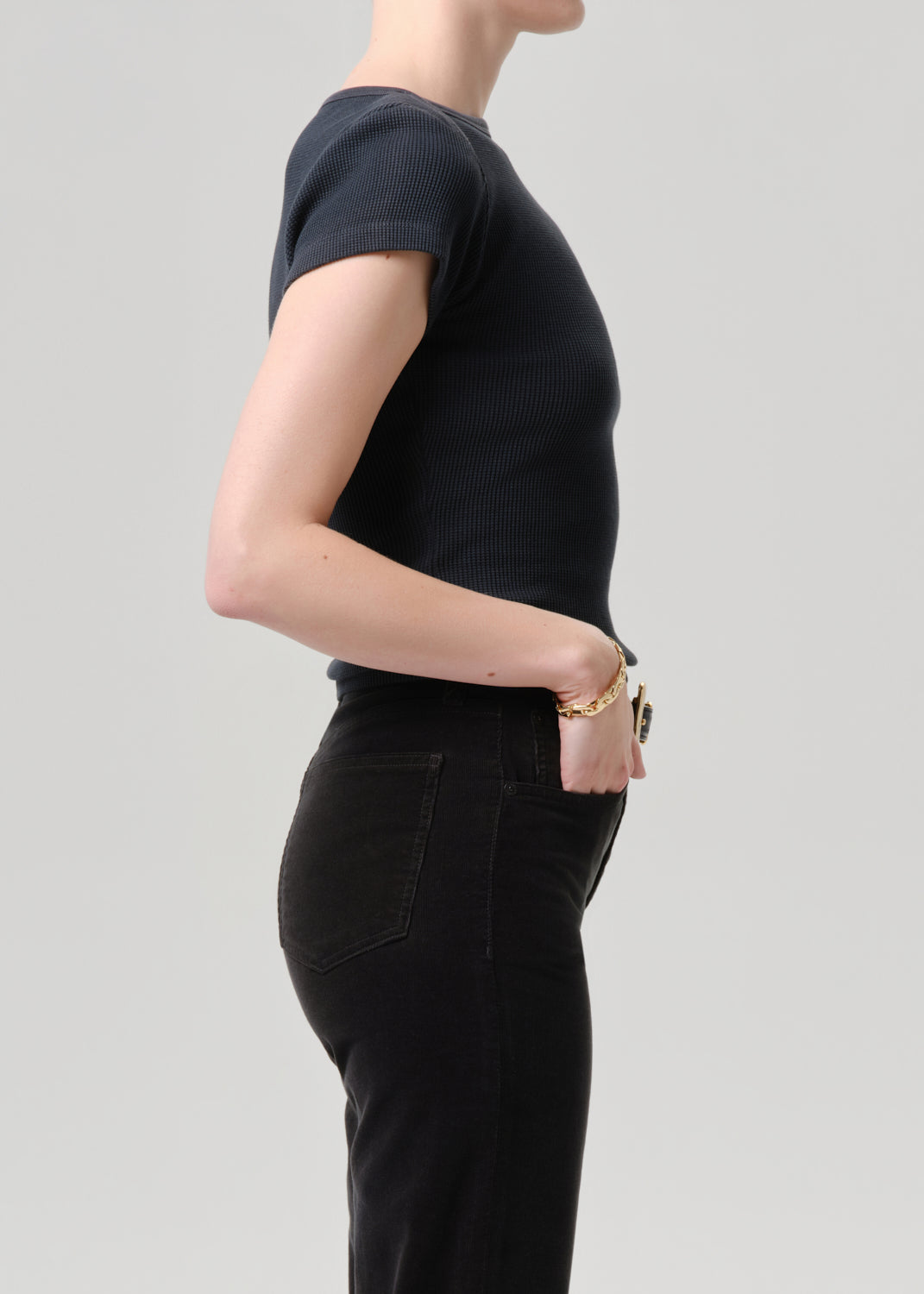 Side profile of a model wearing a black short-sleeve ribbed knit top and black jeans against a grey background.