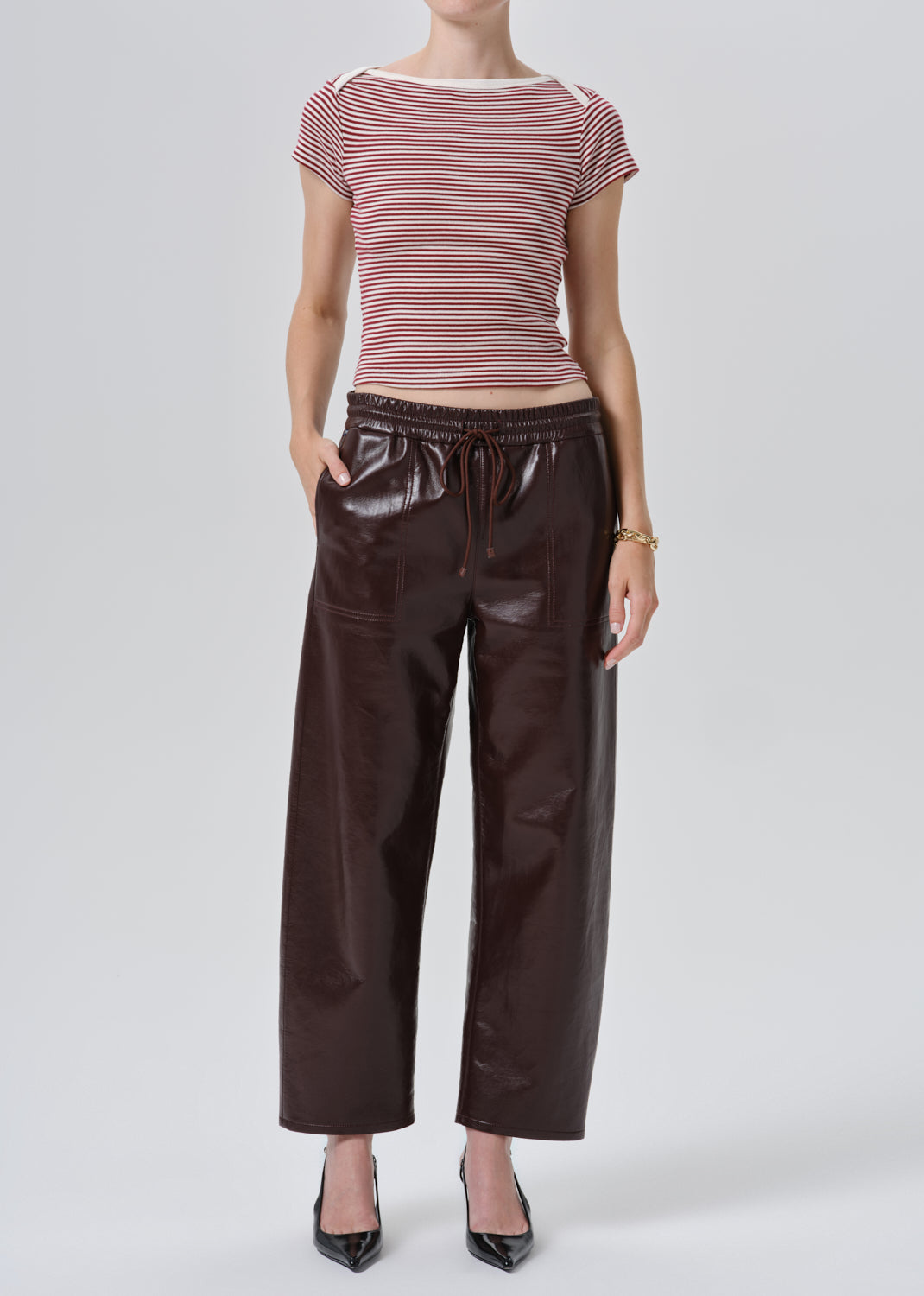 A model wearing a pink and white striped short-sleeve top and dark brown faux-leather wide-leg pants, paired with black pointed-toe heels, against a grey background.