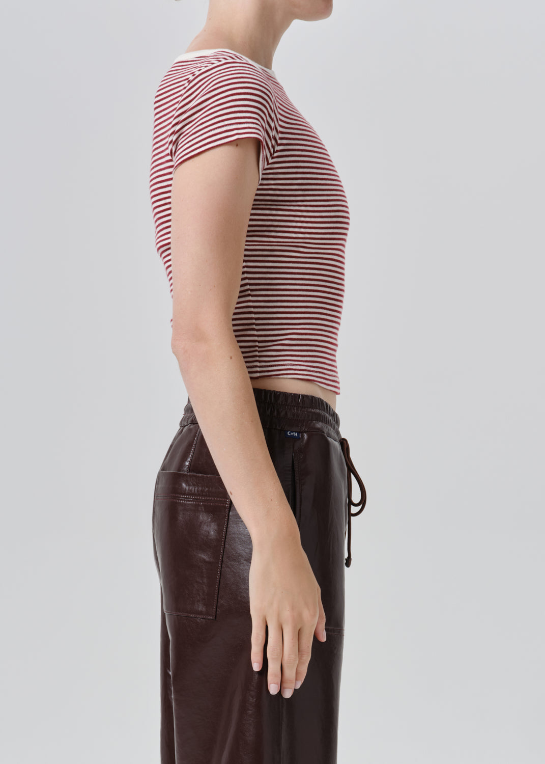 Side profile of a model wearing a short-sleeve cropped top with thin red and white horizontal stripes, paired with dark brown pants, against a grey background.
