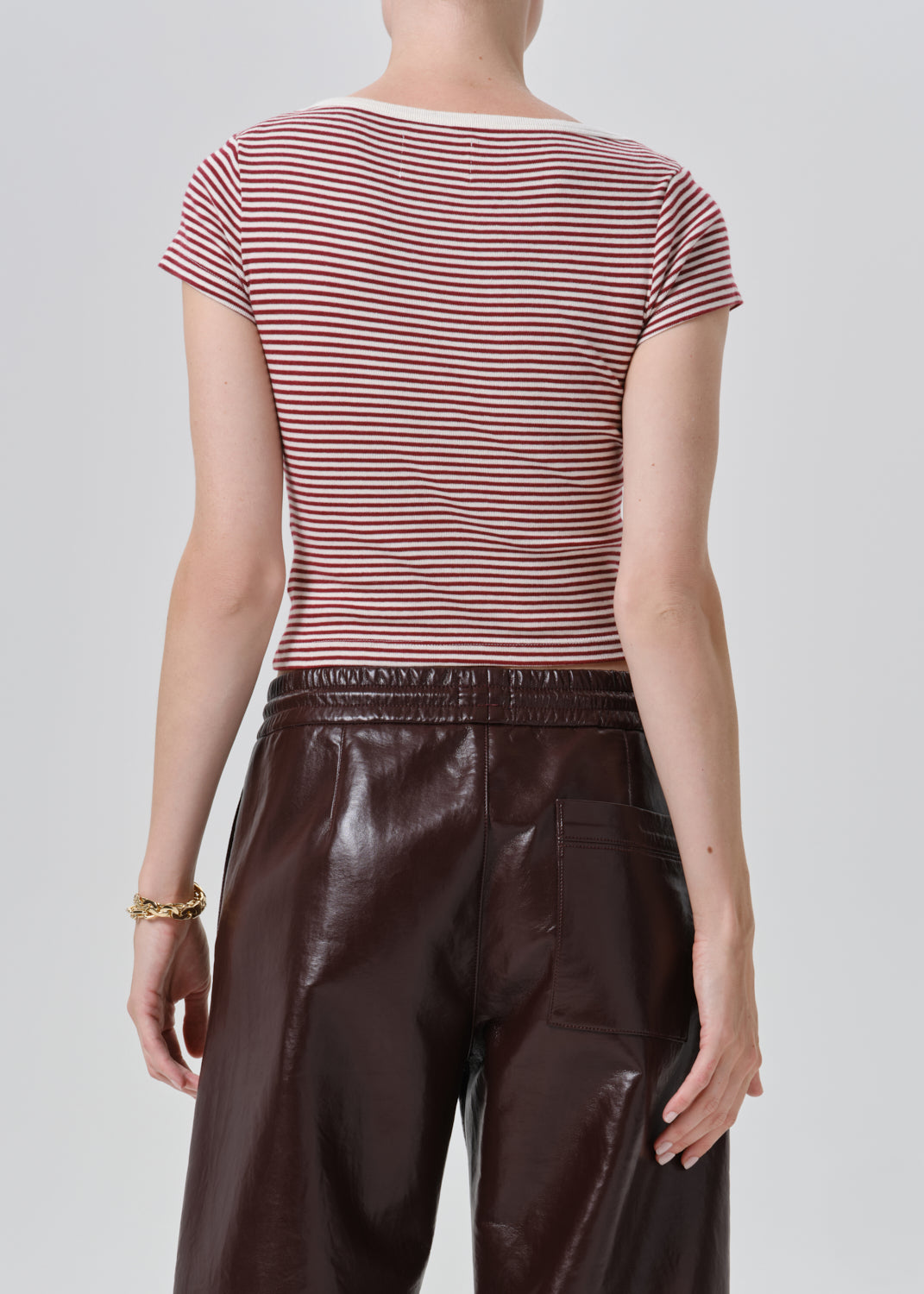 Rear view of a model wearing a short-sleeve crew-neck top with thin red and white horizontal stripes, paired with dark brown faux-leather pants, against a grey background.