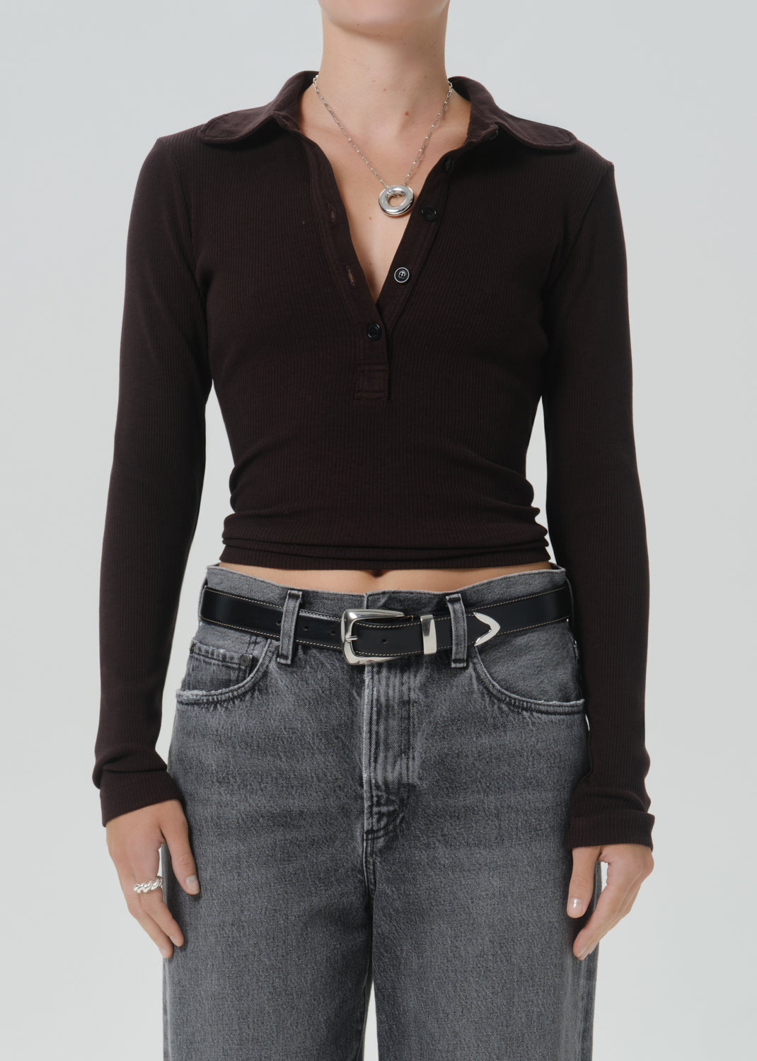 A model wearing a dark brown long-sleeve polo henley top tucked into grey jeans with a black belt, shown from the neck down against a grey background.