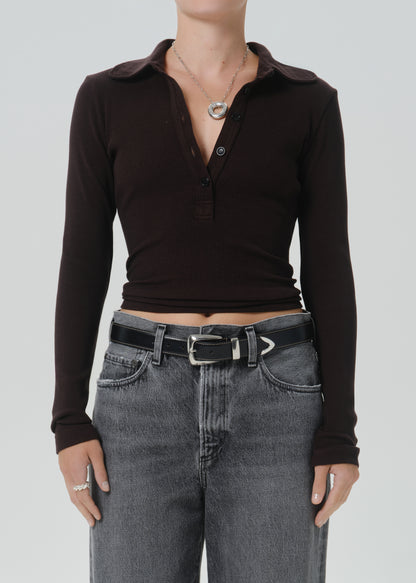 A model wearing a dark brown long-sleeve polo henley top tucked into grey jeans with a black belt, shown from the neck down against a grey background.