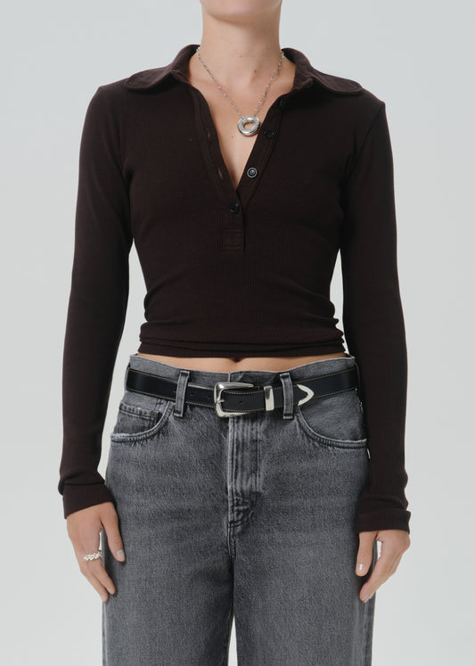A model wearing a dark brown long-sleeve polo henley top tucked into grey jeans with a black belt, shown from the neck down against a grey background.