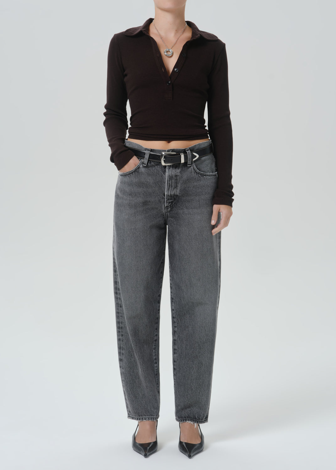 A model wearing a dark brown ribbed long-sleeve polo crop top and high-waisted, wide-leg grey denim jeans with a black belt, paired with black pointed-toe heels, against a grey background.