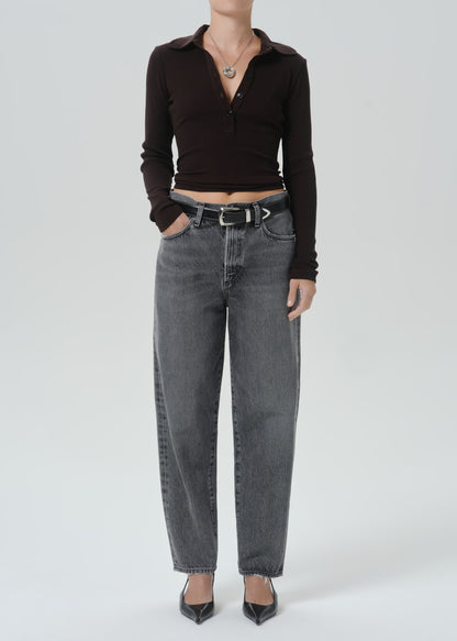 A model wearing a dark brown ribbed long-sleeve polo crop top and high-waisted, wide-leg grey denim jeans with a black belt, paired with black pointed-toe heels, against a grey background.