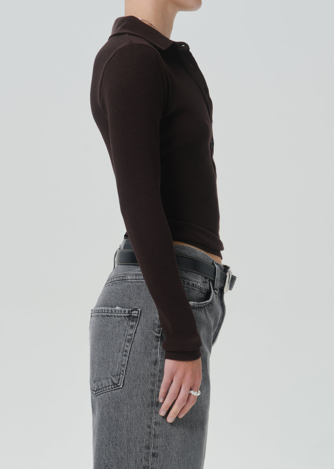 Side profile of a model wearing a dark brown long-sleeve polo shirt tucked into grey denim jeans against a grey background.