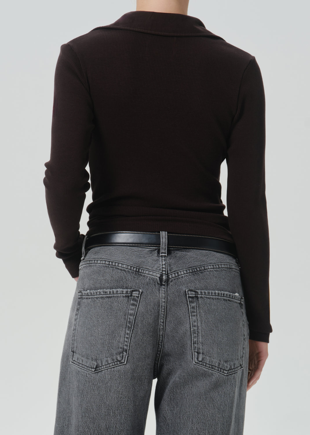 Rear view of a person wearing a dark brown long-sleeve knit top, a black belt, and grey jeans against a grey background.