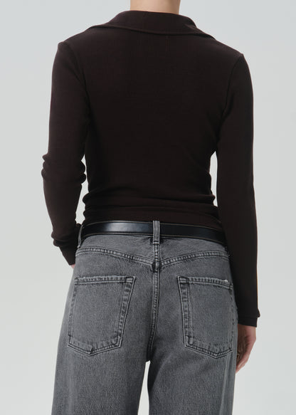 Rear view of a person wearing a dark brown long-sleeve knit top, a black belt, and grey jeans against a grey background.