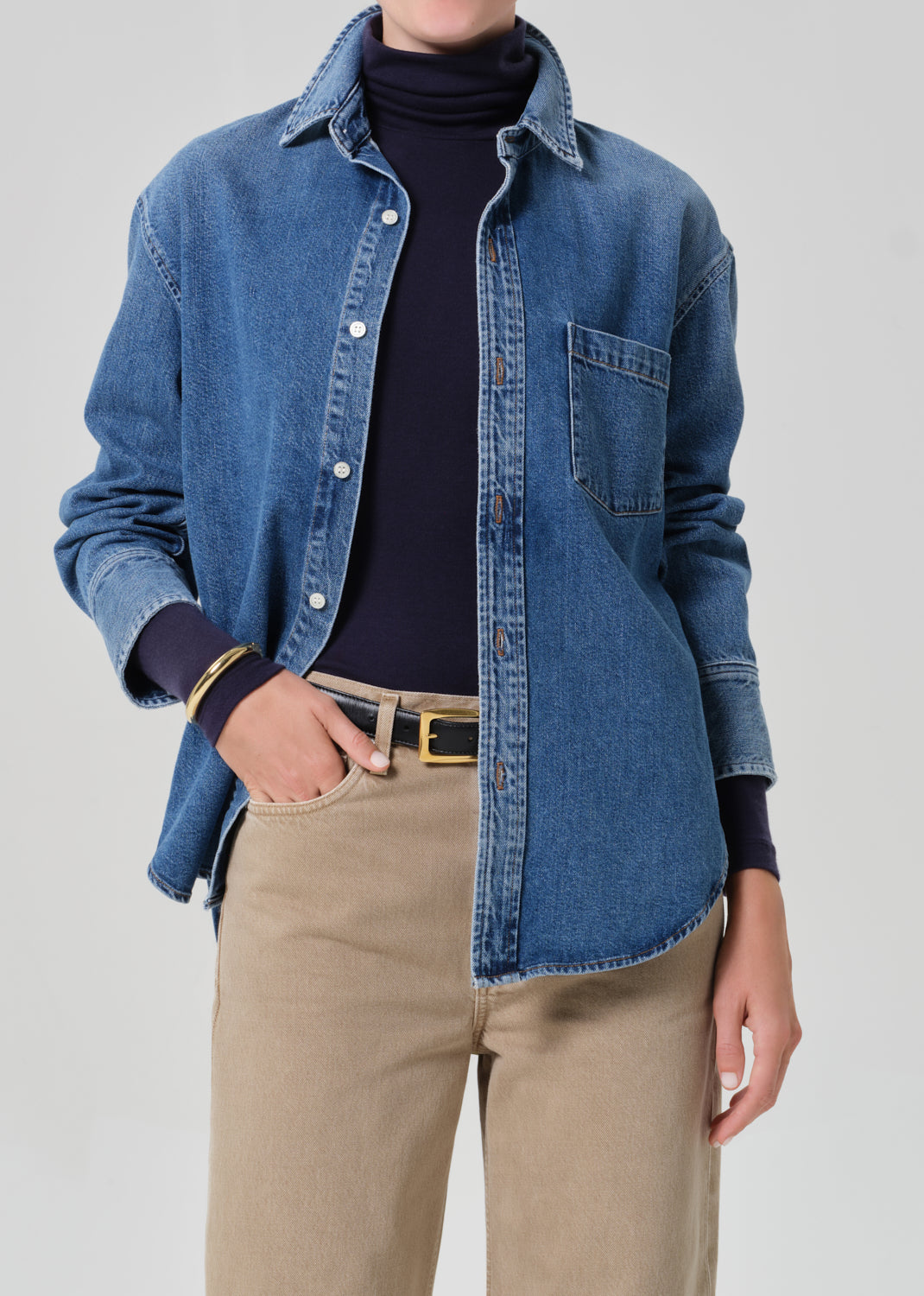 Person wearing a blue denim shirt over a dark turtleneck with beige pants on a gray background