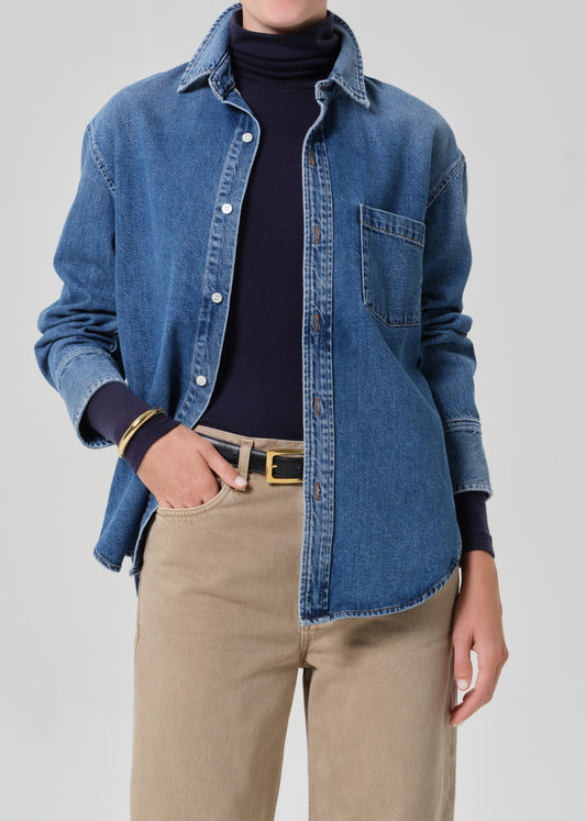 Person wearing a blue denim shirt over a dark turtleneck with beige pants on a gray background