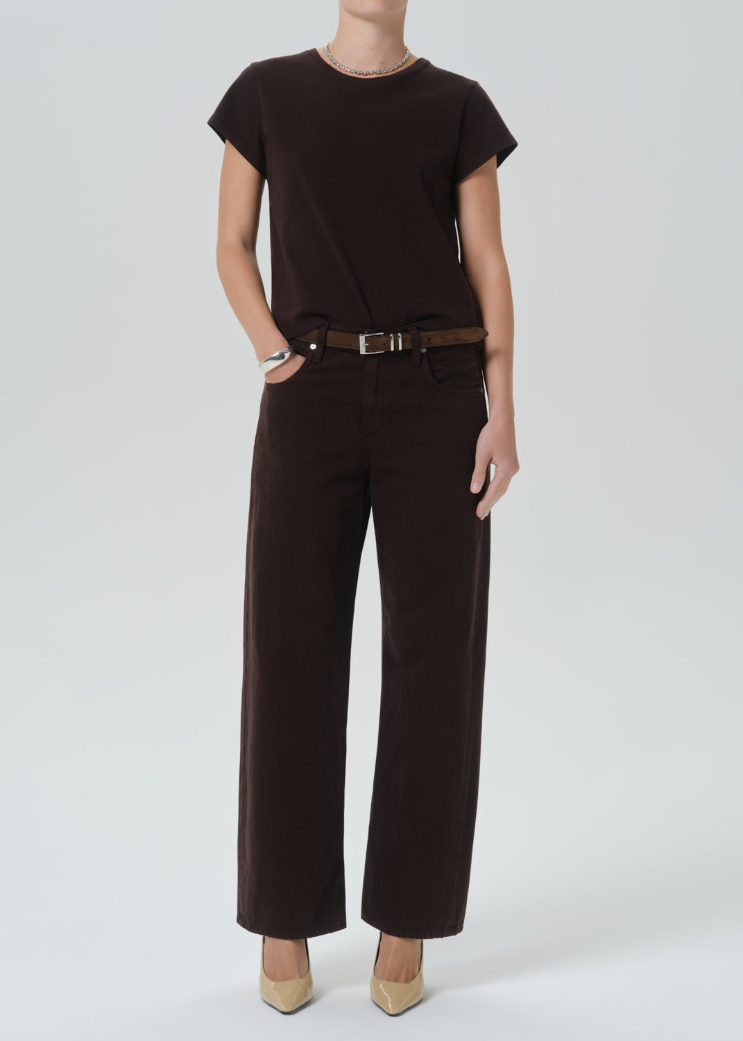 A model wearing a dark brown short-sleeve top and matching dark brown wide-leg trousers with a brown belt, paired with beige pointed-toe heels, against a grey background.