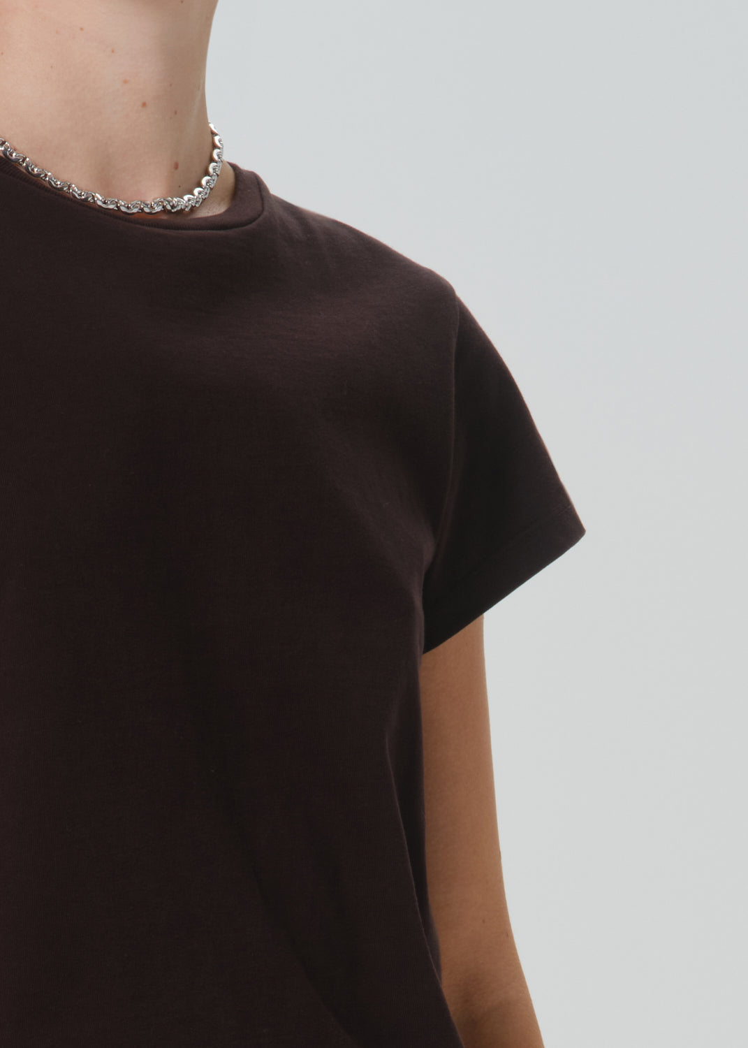Close-up of a person wearing a dark brown short-sleeve crewneck t-shirt and a silver chain necklace, set against a grey background.