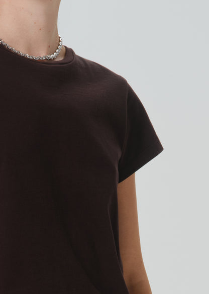 Close-up of a person wearing a dark brown short-sleeve crewneck t-shirt and a silver chain necklace, set against a grey background.