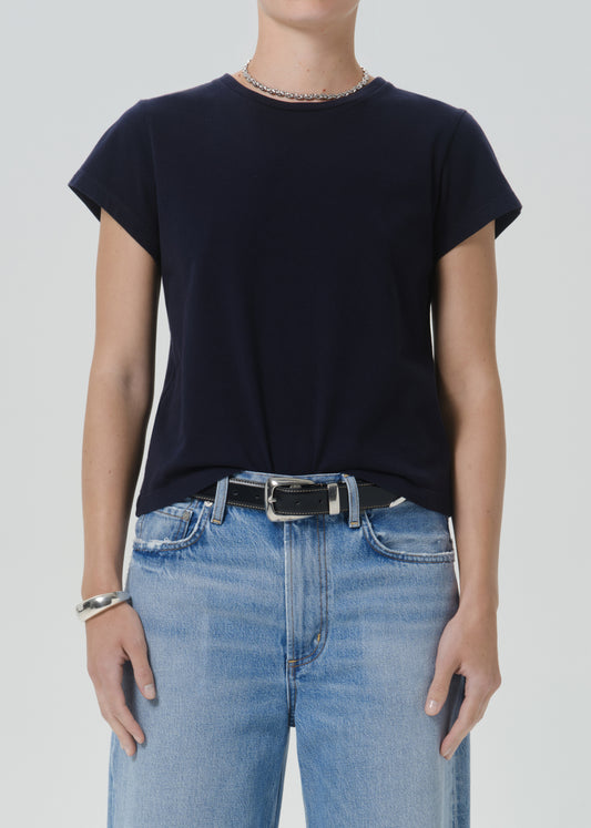 A model wearing a dark navy blue short-sleeve crewneck t-shirt tucked into light-wash blue jeans with a black belt, shown from the neck down against a grey background.